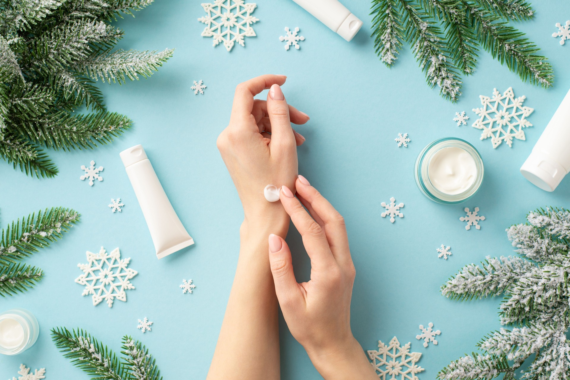 Winter season skin care concept. First person top view photo of woman using hand cream cosmetic jars tubes snowflakes and spruce branches in hoarfrost on isolated pastel blue background with copyspace